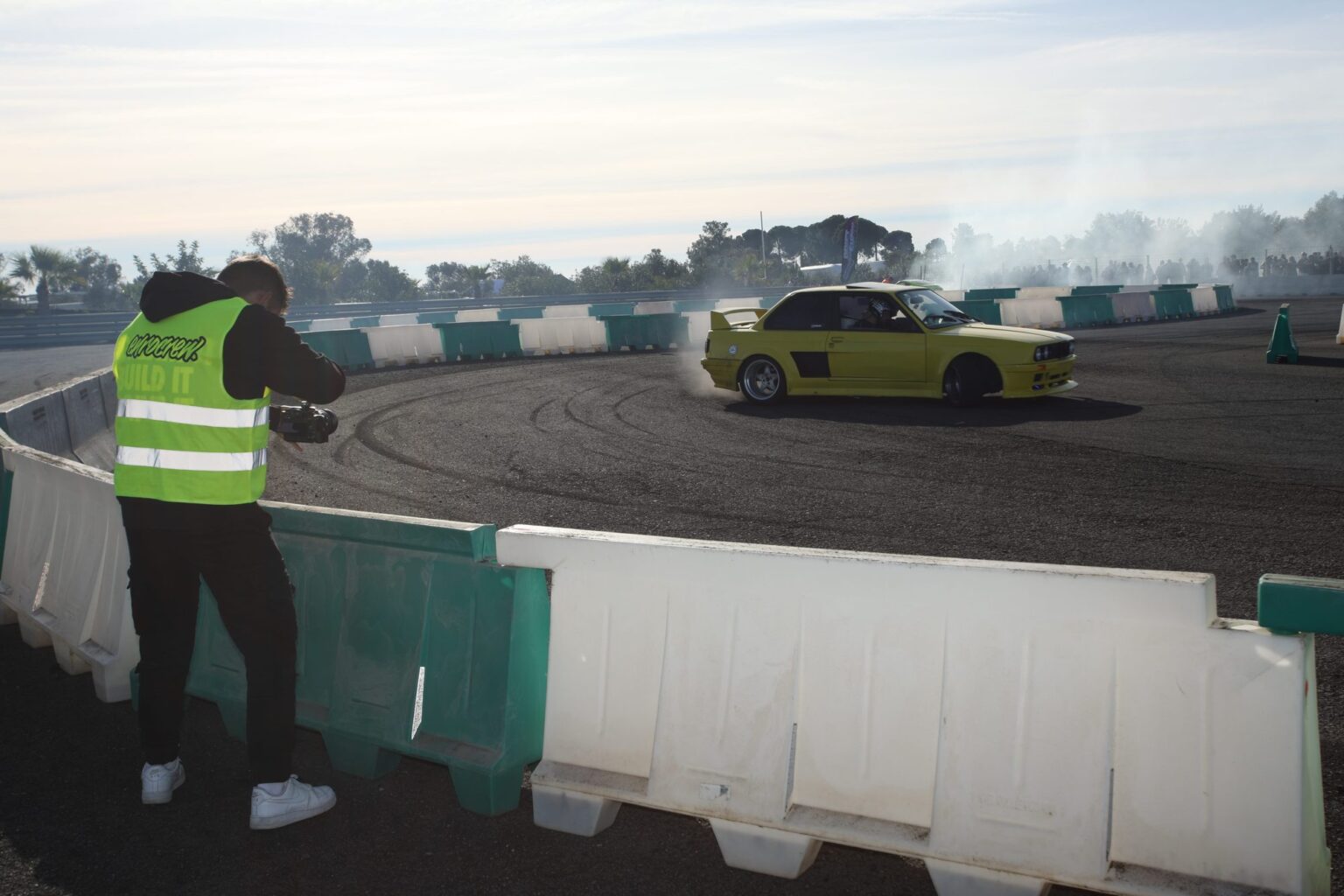 Gonzalo Ochoa grabando con chaleco de prensa junto a la pista mientras un BMW amarillo derrapa entre humo
